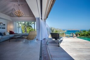 Una sala de estar moderna con un sofá grande, una silla y una mesa de comedor. Se ve el océano y palmeras.