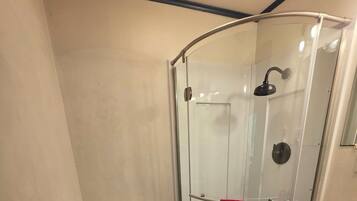 Ducha con mampara de vidrio y puerta curva, inodoro blanco y lavamanos con una toalla roja colgada en la puerta.