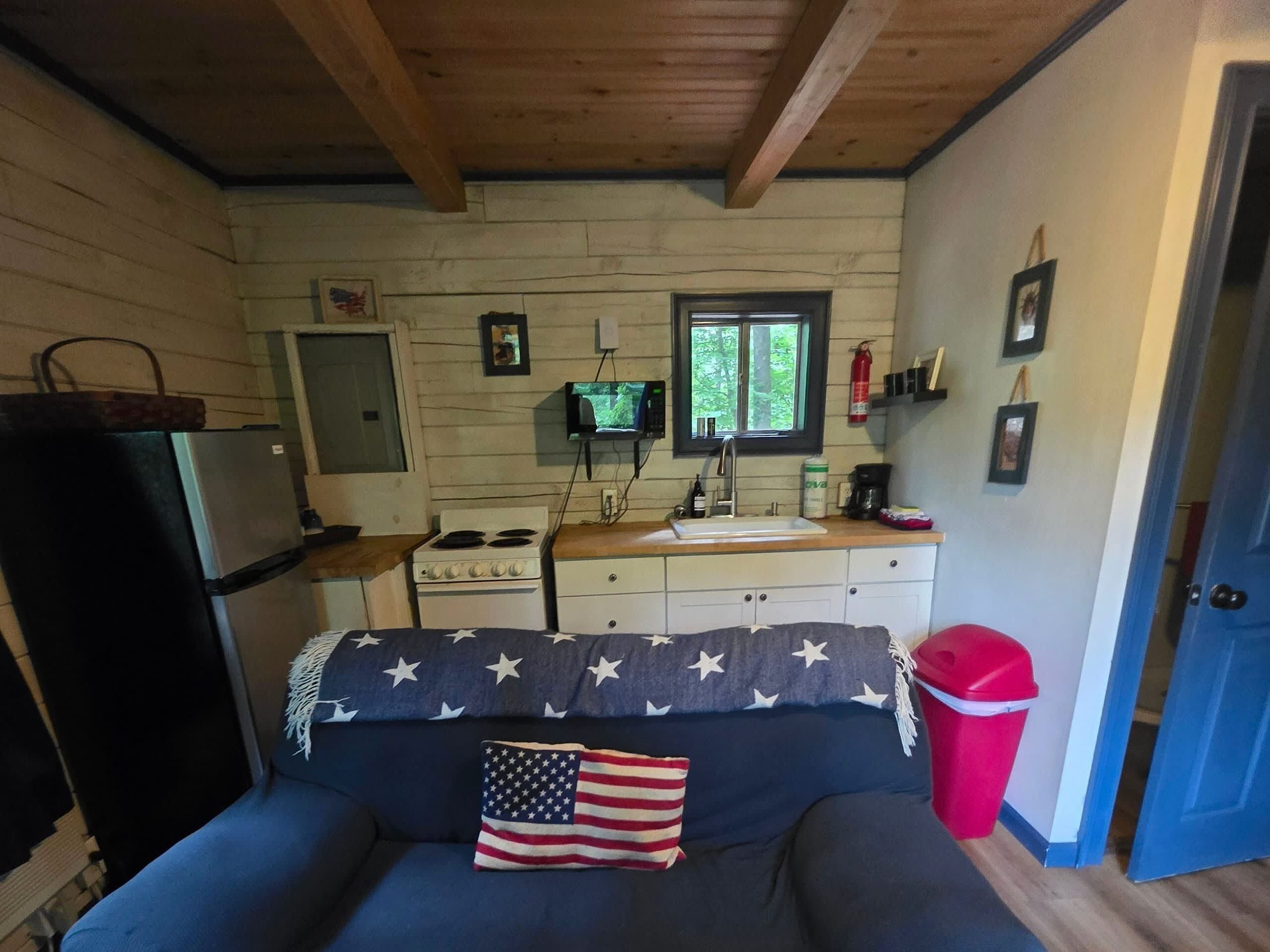 Een compacte keuken met een fornuis, gootsteen en koelkast. Er is een bank met een kussen met de Amerikaanse vlag en een rode prullenbak.