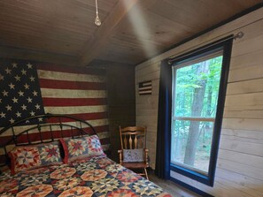 A bedroom with a bed, a chair, a window, and a wall with a flag. - Eagles Nest at Avenwood Cabins (new straitsville)