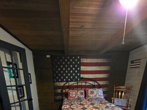 A bedroom with a bed, a wooden ceiling, a wall with an American flag, and a chair with a bicycle pillow.