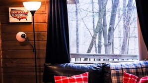 Un accogliente interno di una baita con una parete in legno, una lampada, una panca con cuscini a quadri e una finestra vista alberi.