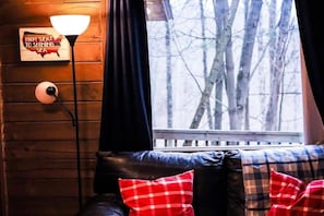 A cozy cabin interior with a wooden wall, a lamp, a bench with plaid cushions, and a window with a view of trees. - Eagles Nest at Avenwood Cabins (new straitsville)
