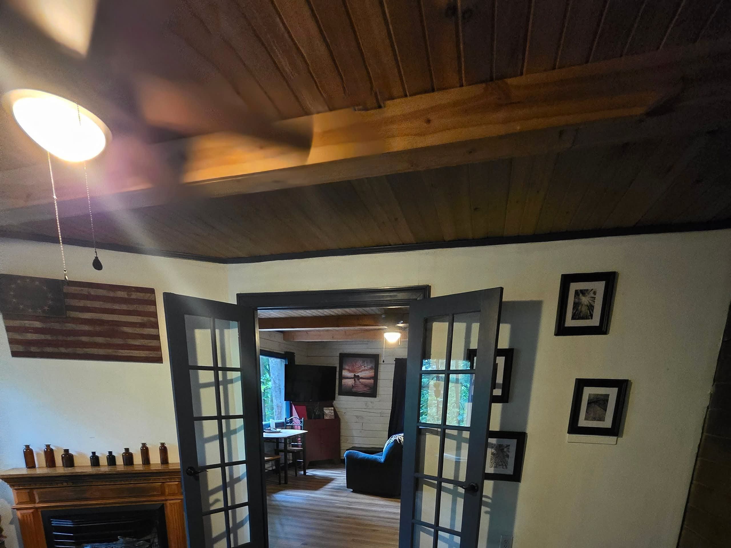 A room with a wooden ceiling, a fireplace, a dining area, and a view through a doorway.