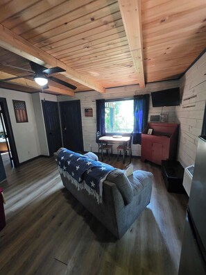 Ein gemütliches Blockhäuschen im Inneren mit Holzbalkendecke, einem Esstisch, einer Couch und einem Fernseher.