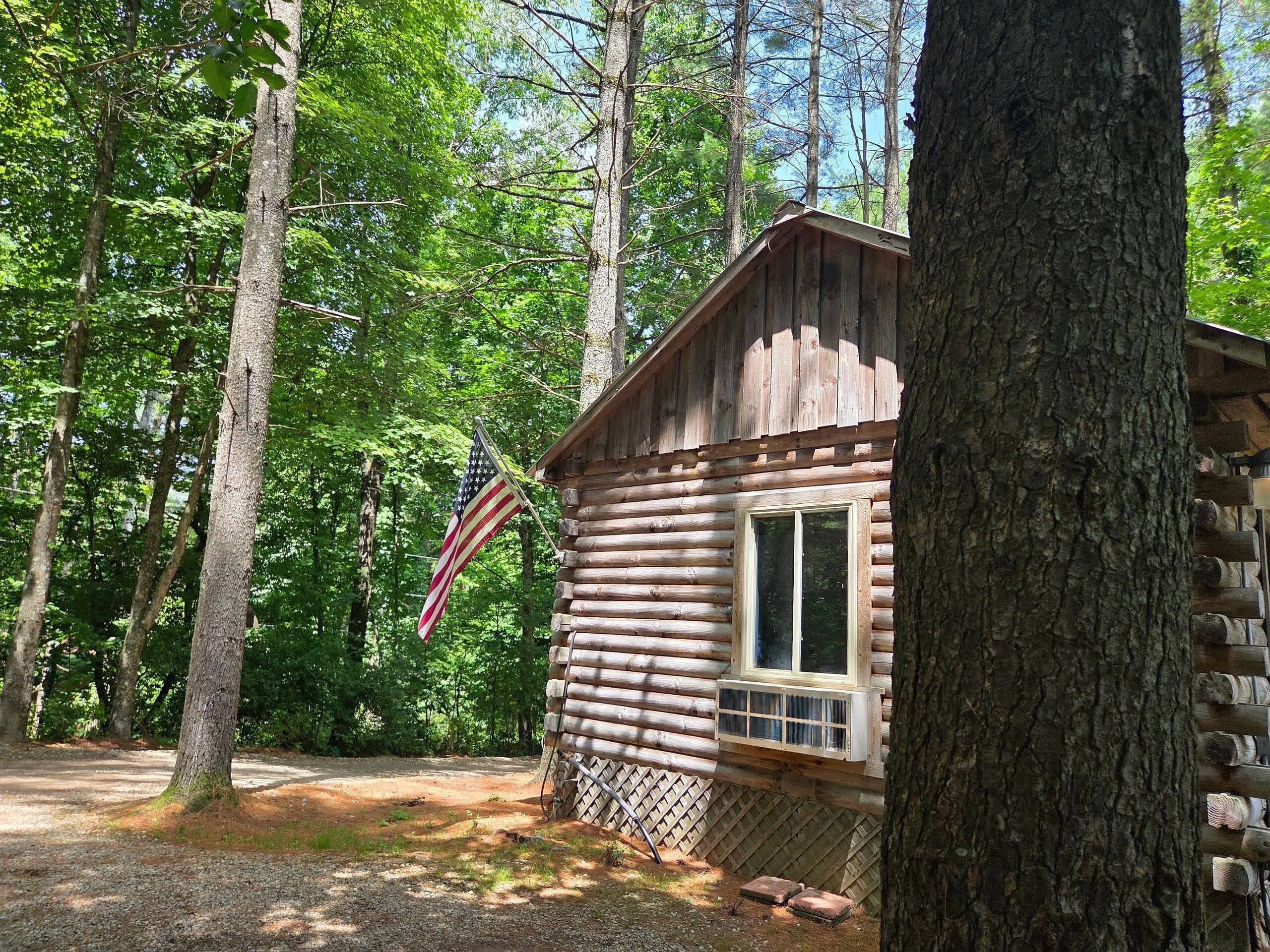 A rustic log cabin with a window and an American flag, surrounded by trees.