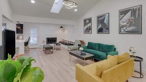 Uma sala de estar moderna com sofá verde, poltrona amarela e lareira. Há quadros nas paredes e um clarabóia acima.