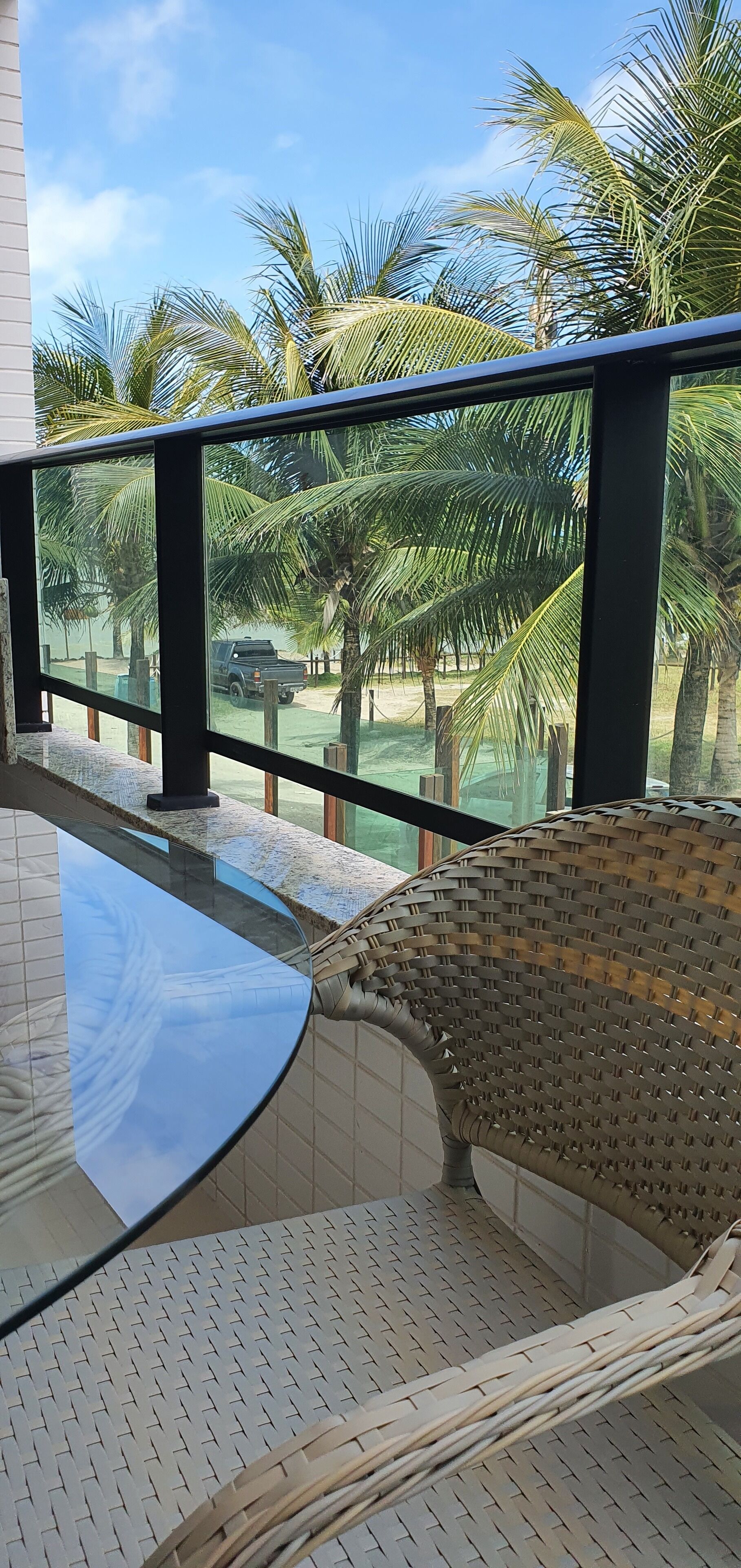 A balcony with a glass table and wicker furniture, overlooking a palm tree-lined area with a parked vehicle.