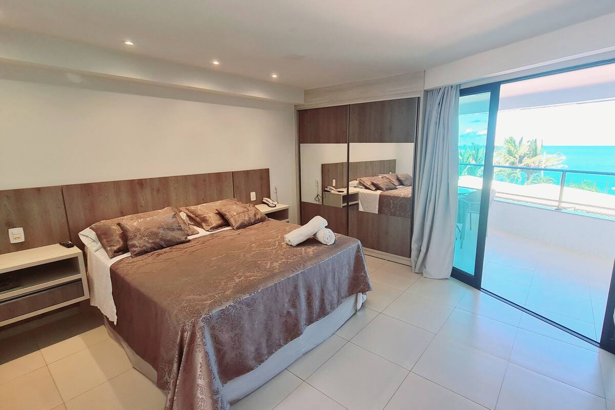 A modern hotel room with a large bed, a nightstand, a mirror, and a sliding glass door leading to a balcony with a view of the sea.
