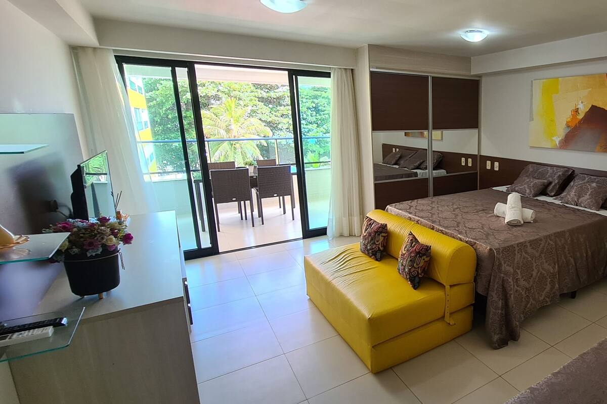 A modern hotel room with a bed, a yellow sofa, a balcony with a table and chairs, and a painting on the wall.