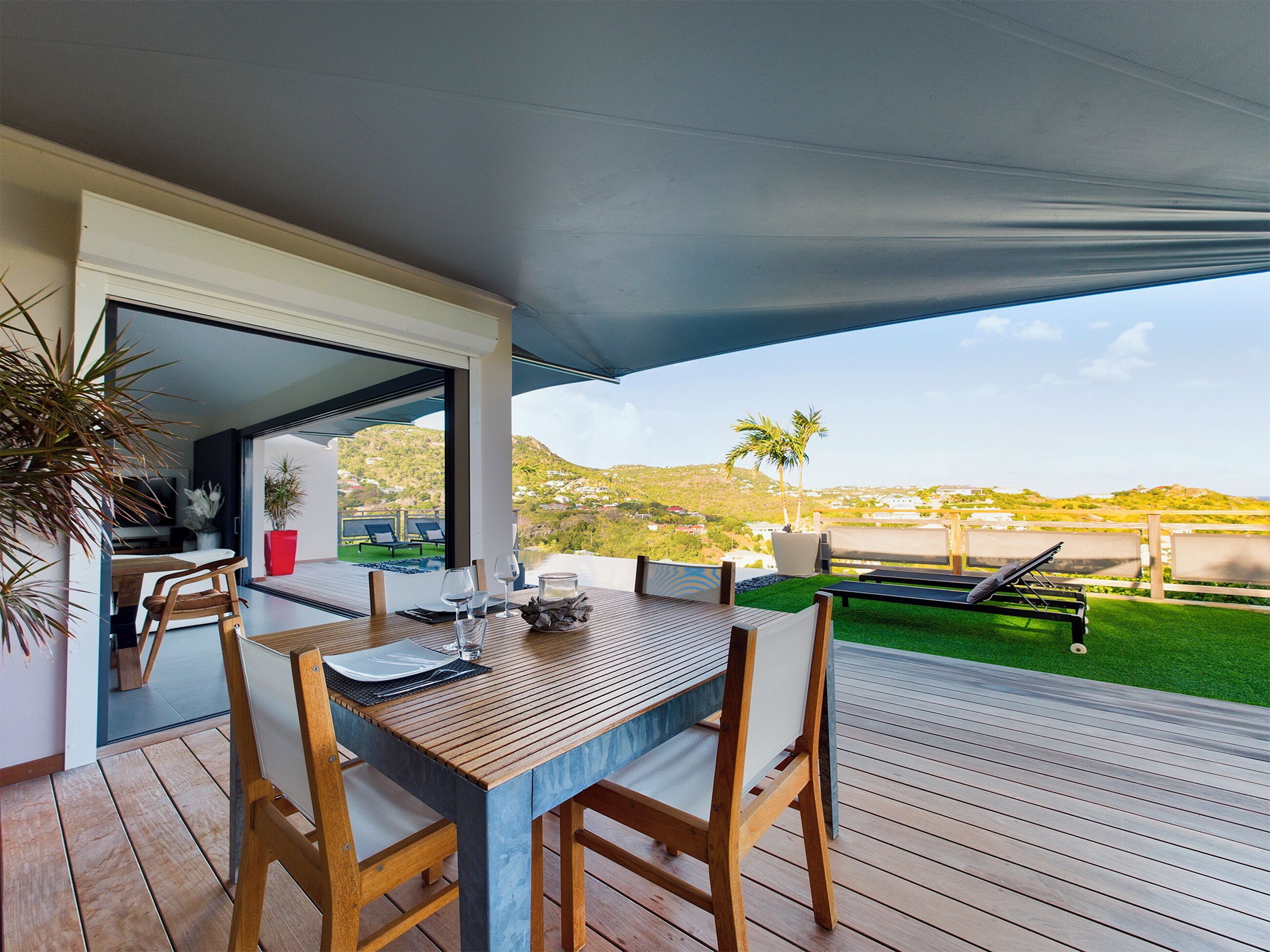 Un espace de restauration en plein air avec une table et des chaises en bois, un patio couvert et une vue sur un paysage verdoyant et des collines en arrière-plan.