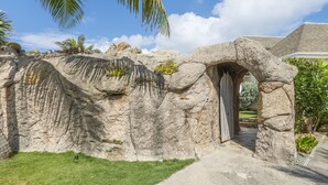 Un arco de piedra que da acceso a un edificio, rodeado de un césped cuidado y palmeras.