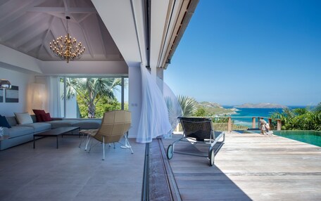 A modern living room with a large sofa, a chair, and a dining table. There is a view of the ocean and palm trees.