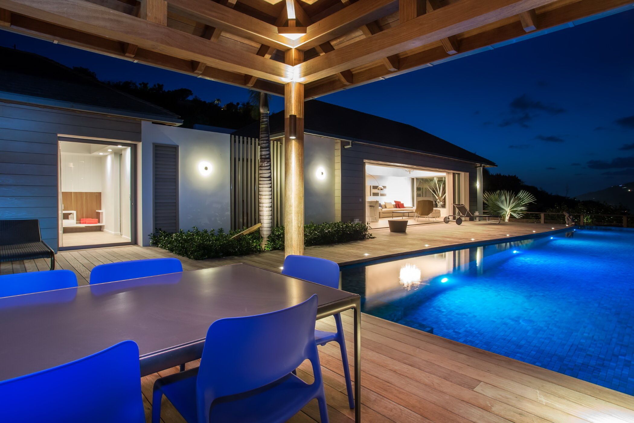 A modern outdoor pool area with a wooden deck, a dining table with chairs, and a well-lit house in the background.