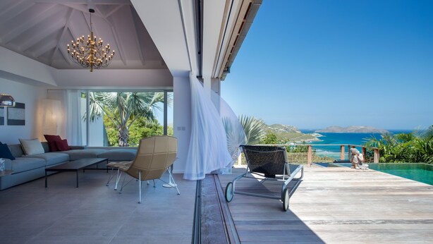Ein modernes Wohnzimmer mit einer großen Couch, einem Sessel und einem Esstisch. Blick aufs Meer und auf Palmen.