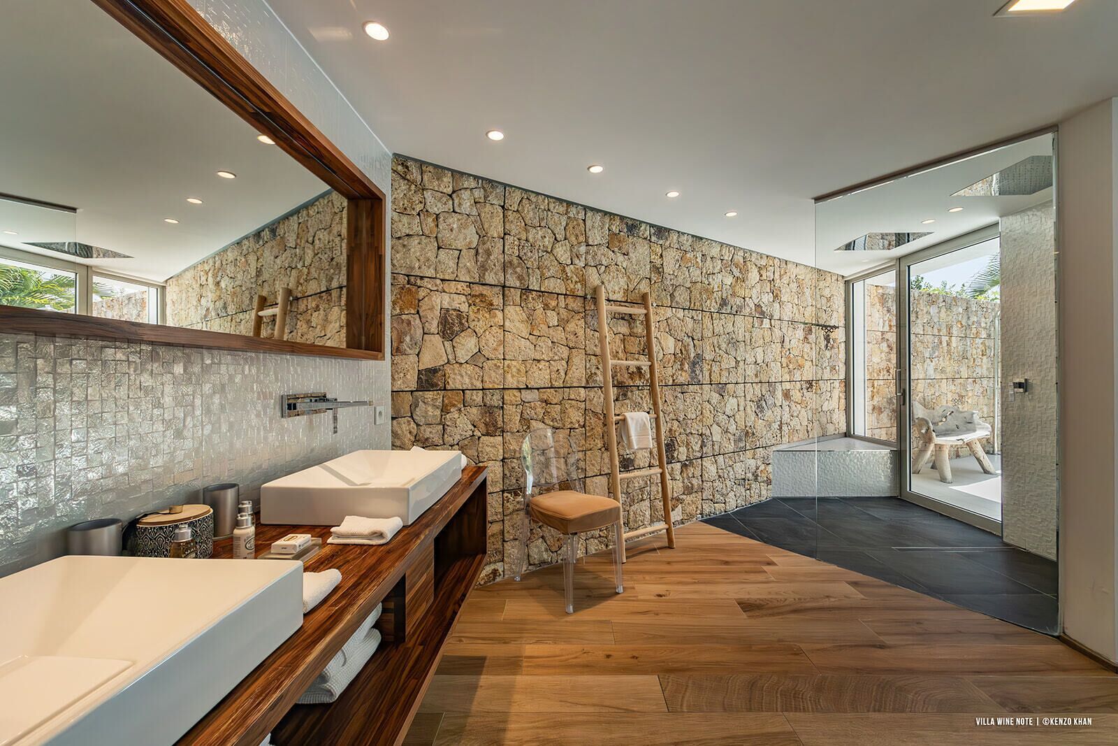 A modern bathroom with a stone wall, two sinks, a wooden vanity, and a chair.
