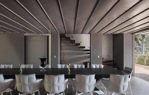 A modern dining area with a long table, white chairs, and a staircase leading to an outdoor area. - Villa Dunes  | Near Ocean - Located in  Magnificent Salines with Private Pool (Gustavia)