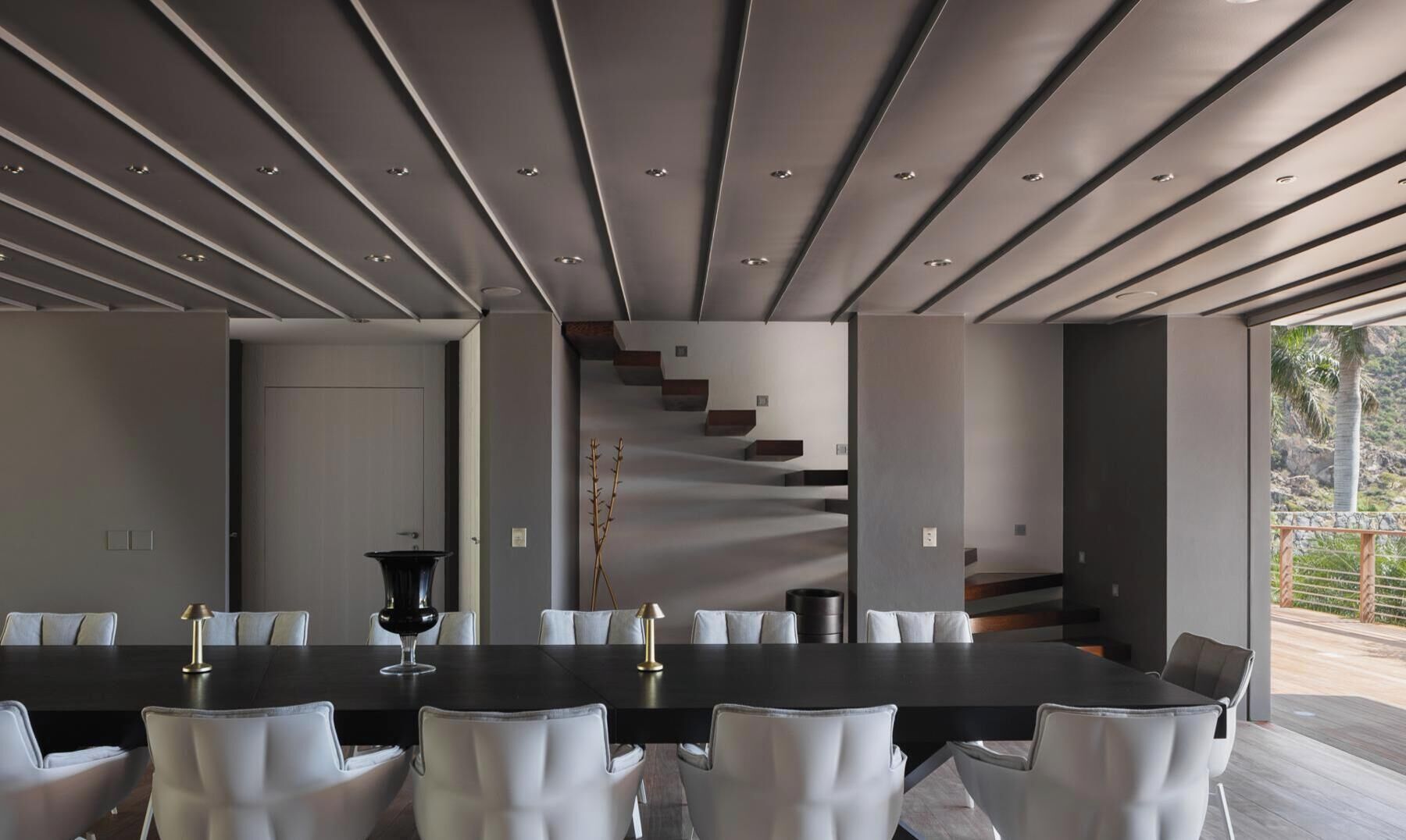 Un espace de salle à manger moderne, avec une longue table, des chaises blanches et un escalier menant à un espace extérieur.