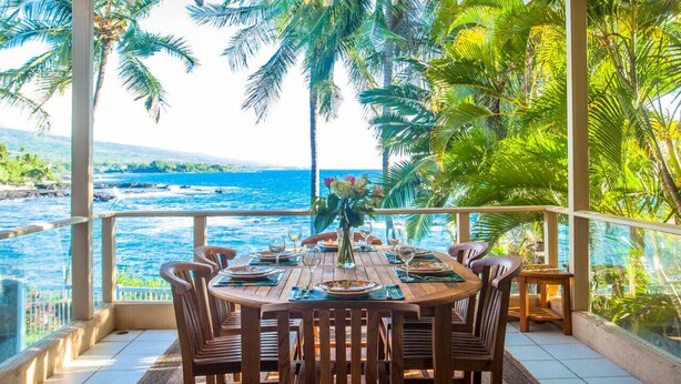 A dining area with a table set for four, overlooking a coastal view with palm trees and the ocean. - Oceanfront Luxury at Alii Point | Private Pool, Panoramic Views & Tropical Serenity (Kailua-Kona)