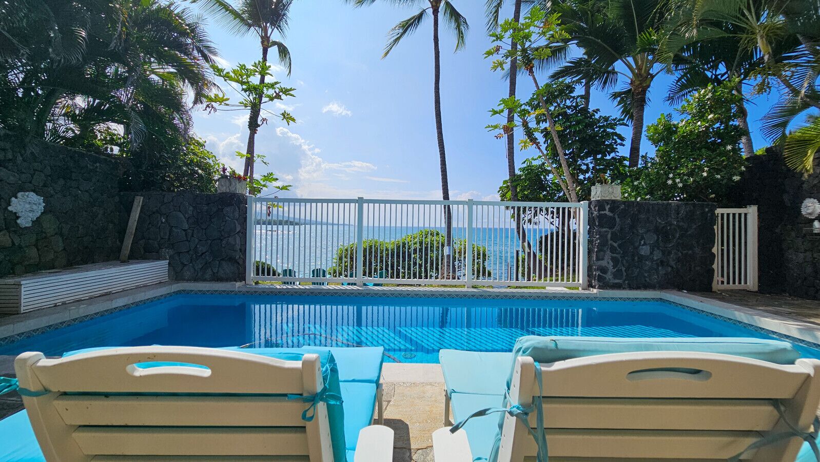 A pool with two lounge chairs, a stone wall, and a view of the ocean.