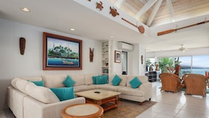 A spacious living room with a large sectional sofa, a small round table, and wicker chairs. There is a large painting of a coastal scene on the wall, and a view of the sea through large windows. - Oceanfront Luxury at Alii Point | Private Pool, Panoramic Views & Tropical Serenity (Kailua-Kona)