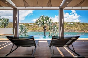 Zona de piscina con dos sillones reclinables, una terraza de madera y vista a una colina con palmeras y una masa de agua.
