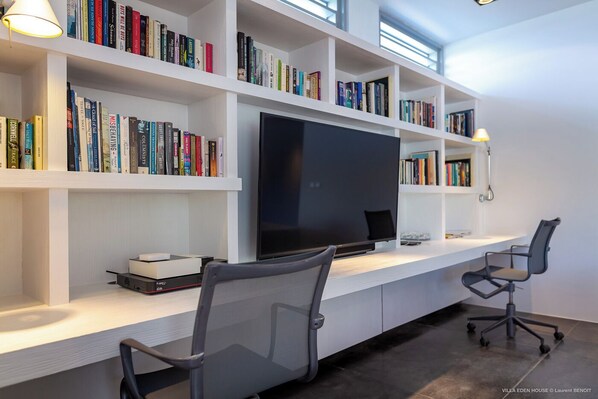 Una oficina en casa moderna con una televisión de pantalla plana grande, estanterías blancas con libros, dos sillas de escritorio y un escritorio.