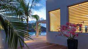 Une villa moderne dotée d’une terrasse en bois, de grandes fenêtres équipées de stores horizontaux et offrant une vue sur la mer.