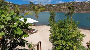 Une vue sur la plage, avec un parasol blanc et une chaise longue, le tout entouré de verdure et donnant sur une étendue d’eau calme.
