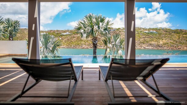 Un espace piscine comprenant deux chaises longues, une terrasse en bois et une vue sur une colline parsemée de palmiers et bordée d’un plan d’eau.