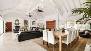 A spacious living room with a large sofa, dining table, and chairs. There is a fireplace, ceiling fans, and large windows. - Villa Petite Plage 4  | Beach Front - Located in  Stunning Grand Case with Private Pool (Grand-Case)
