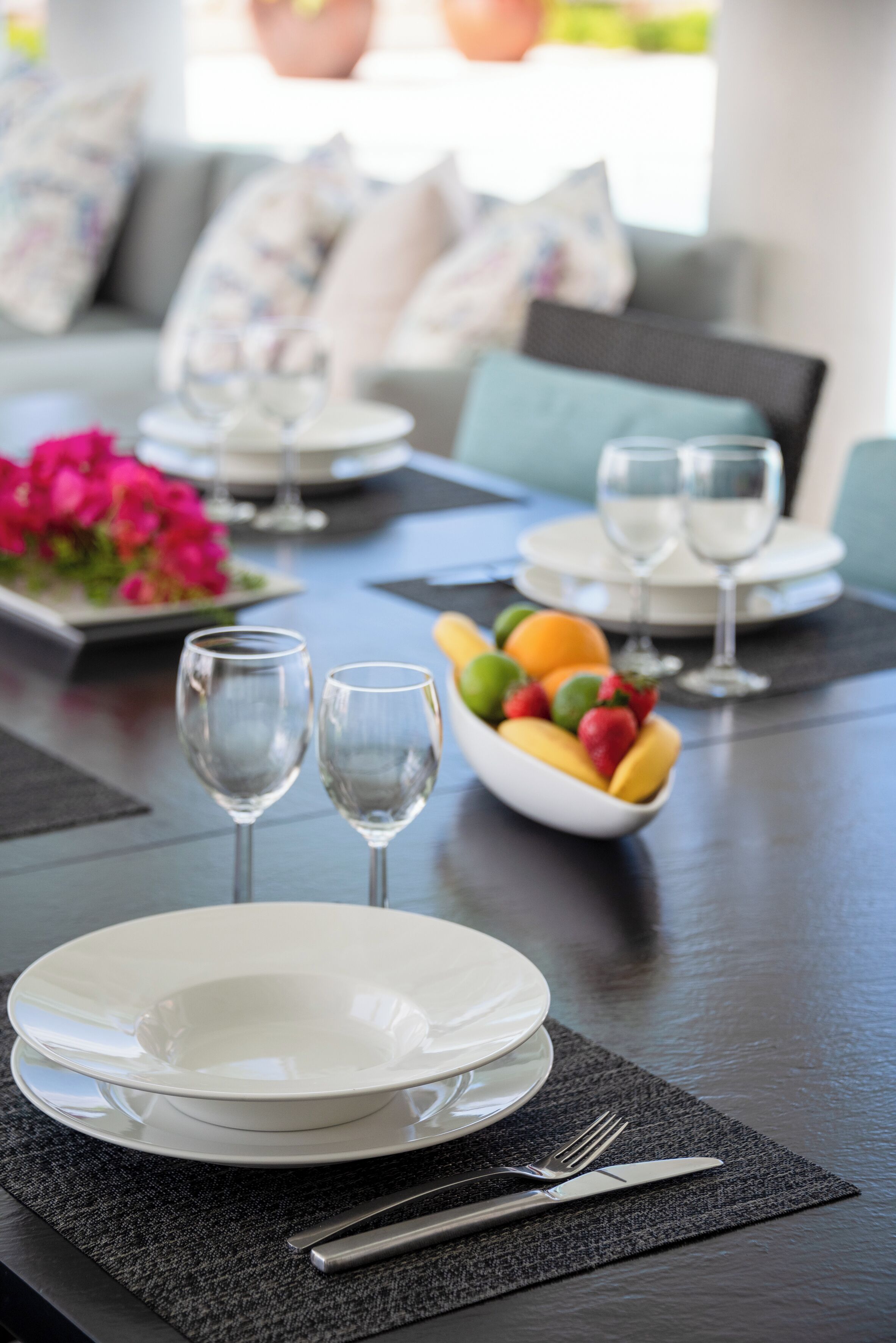 Une table à manger dressée avec des assiettes, des verres et des couverts, avec un saladier de fruits et un vase de fleurs en arrière-plan.
