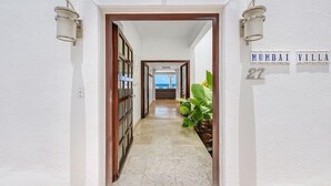 Een hal die leidt naar een kamer met uitzicht op zee, een bordje met 'Mumbai Villa 27' en een bloempot aan de rechterkant.