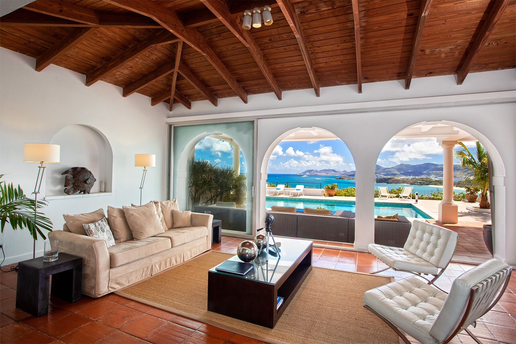A spacious living area with a view of a pool and ocean, featuring a wooden ceiling, a sofa, a coffee table, and a chair.