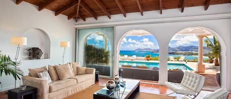 A spacious living area with a view of a pool and ocean, featuring a wooden ceiling, a sofa, a coffee table, and a chair.