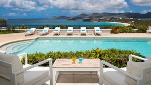 Un espace aménagé au bord de la piscine, avec des chaises longues et une table, offrant une vue sur une plage et des montagnes.
