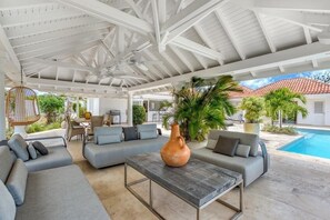 A covered outdoor seating area with a pool, grey sofas, a table, and a hanging chair.