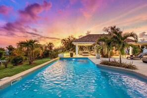 A swimming pool with a covered area and lounge chairs, surrounded by palm trees and a sunset sky.