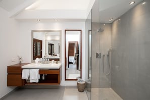 Un baño moderno con cabina de ducha acristalada, tocador con espejo y vista a un dormitorio por una puerta abierta.