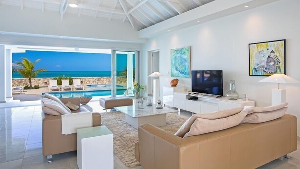 A modern living room with a large TV, comfortable seating, and a view of a pool and ocean.