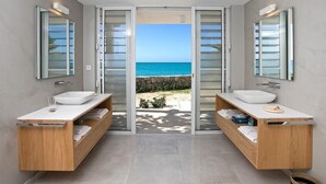 A modern bathroom with two sinks, wooden cabinets, and a view of the sea through glass doors.