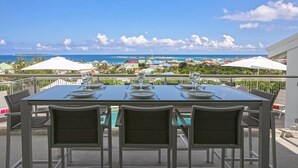 Et spisebord med stole på en udendørs terrasse med udsigt til havet og strandparasoller.