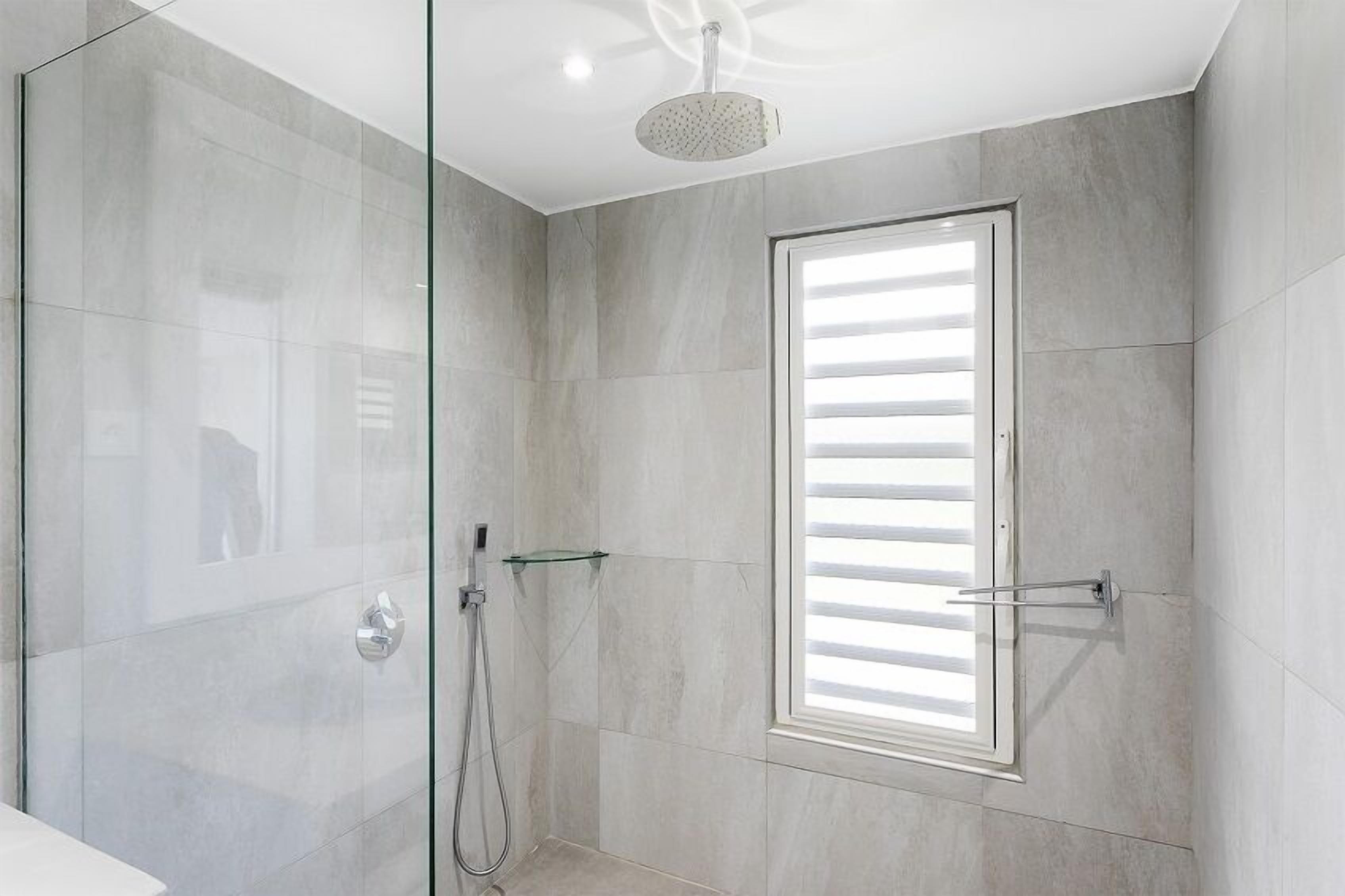 Een moderne badkamer met een douchecabine van glas, een glazen plank en een raam met zonwering.