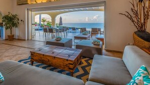 A spacious living area with a large sofa, a wooden coffee table, and a view of the ocean through an open patio. - Villa Happy Bay  | Beach View - Located in  Beautiful Happy Bay with Private Pool (Saint Martin)