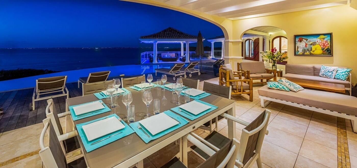 A dining area with a view of the ocean, a covered outdoor seating area, and a sofa with cushions.