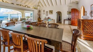 A spacious dining area with a large wooden table and chairs, a view of the sea, and a cozy seating area with a fireplace. - Villa Happy Bay  | Beach View - Located in  Beautiful Happy Bay with Private Pool (Saint Martin)