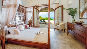 A bedroom with a four-poster bed, a wooden dresser, a chair, and a view of the ocean through an open door. - Villa Happy Bay  | Beach View - Located in  Beautiful Happy Bay with Private Pool (Saint Martin)
