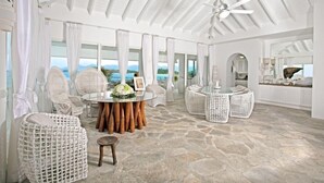 A spacious room with a stone floor, a round dining table with chairs, wicker furniture, and large windows offering a view of the mountains.