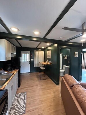 A compact kitchen with a sink, stove, and countertop, a dining area with a table and chairs, and a living area with a sofa. - The Old Moonshiners at Avenwood Cabins (new straitsville)