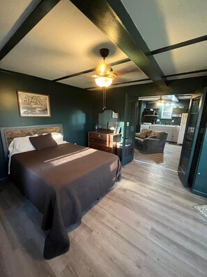 Ein Schlafzimmer mit Bett, Kommode, Nachttisch, Deckenventilator und Blick in einen Küchenbereich.